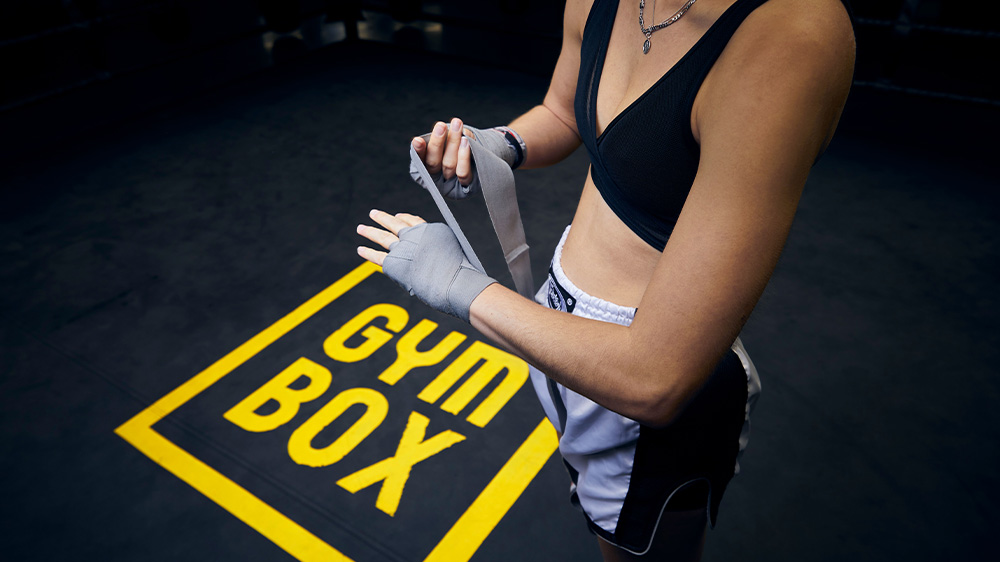Boxing Rookie, woman putting on boxing gear including full Gymbox logo on floor.